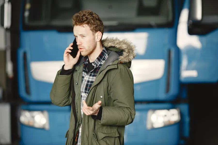 A service technician in uniform taking a phone call next to a work van parked in a driveway