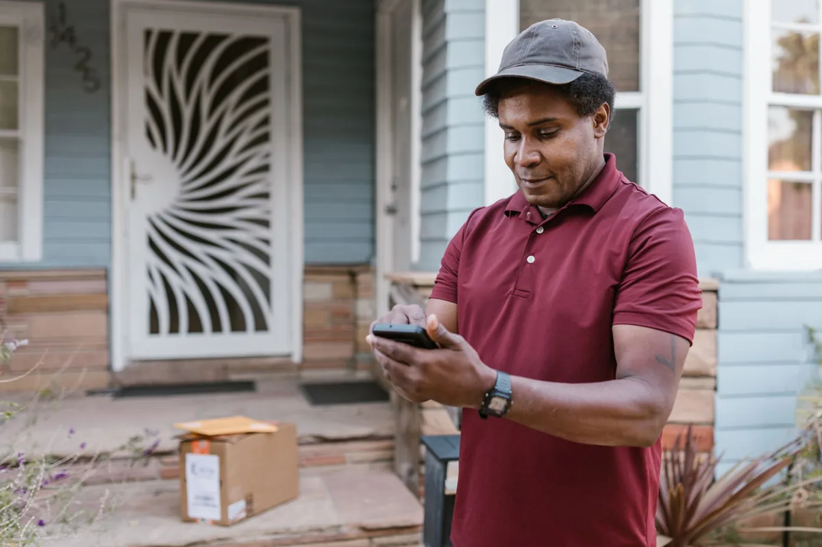 A uniformed home-service technician taking a phone call on a homeowner's front porch next to a parked work van