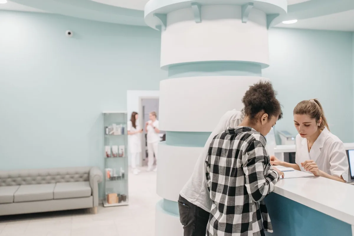 A front-desk receptionist welcoming a patient who arrived on time at a clean, well-lit clinic reception desk