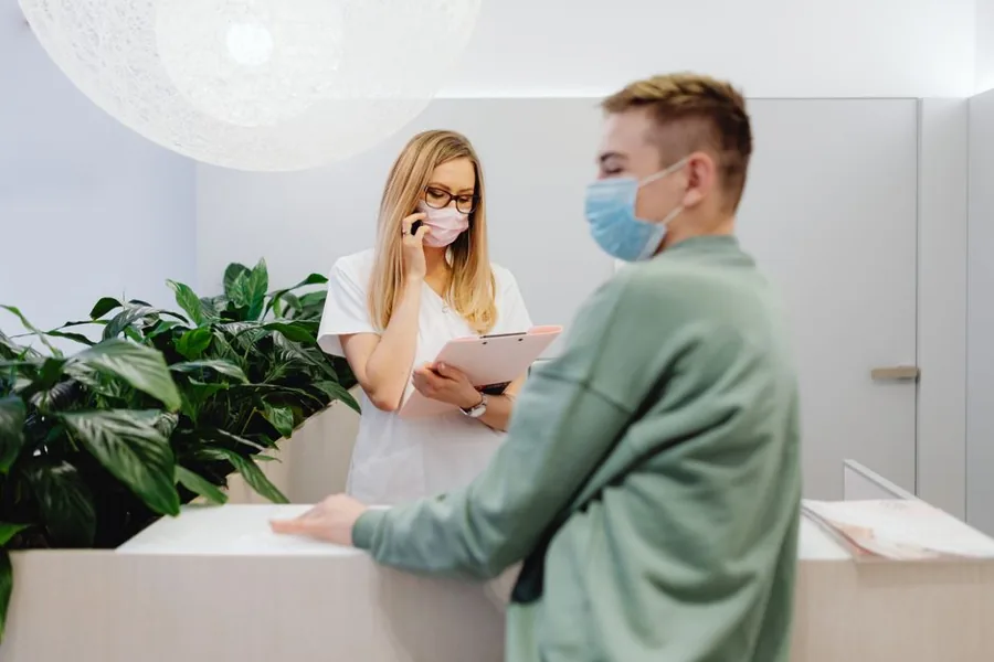 A front-desk receptionist welcoming a patient who arrived on time at a clean, well-lit clinic reception desk